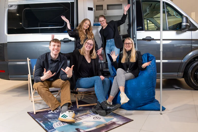 Gruppe junger Erwachsener lächelt und posiert fröhlich vor einem neuen Van in einem Autohaus.